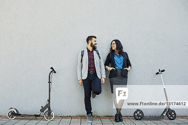 Smiling businessman and businesswoman with scooters leaning against a wall