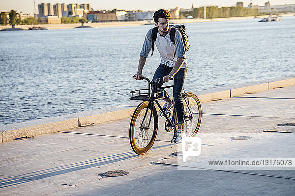 Young man with backpack riding bike on waterfront promenade at the riverside