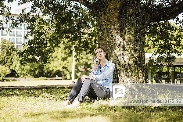 Frau sitzt im Stadtpark und lehnt an einen Baum und telefoniert