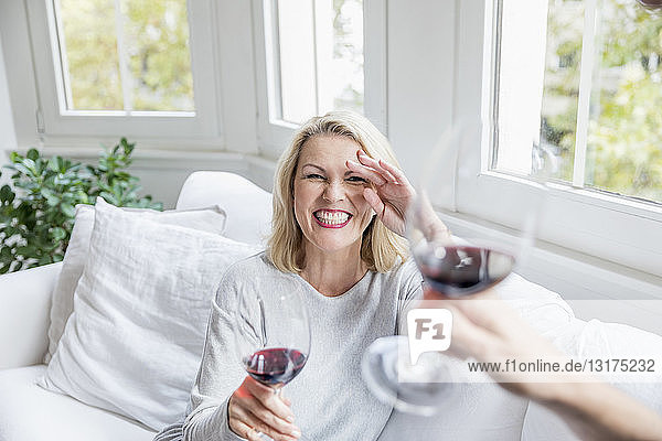 Porträt einer lachenden  blonden  reifen Frau  die zu Hause mit Rotwein anstößt