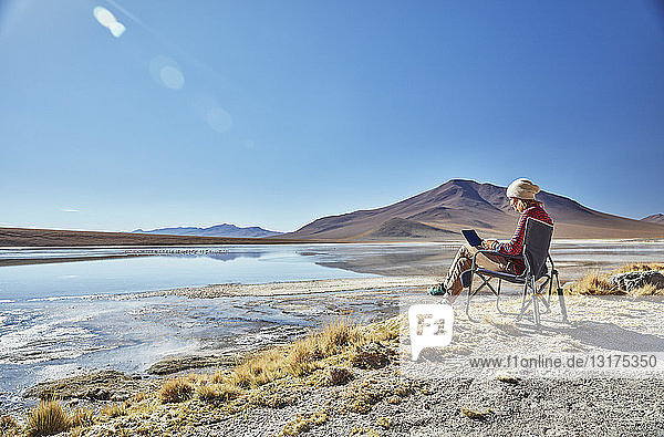 Bolivien  Laguna Colorada  Frau sitzt auf einem Campingstuhl am Seeufer und benutzt eine Tablette