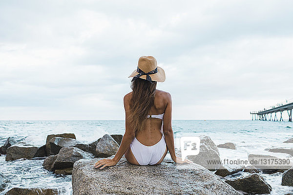 Rückansicht einer jungen Frau in Badeanzug und Hut  die auf einem Felsen im Meer sitzt