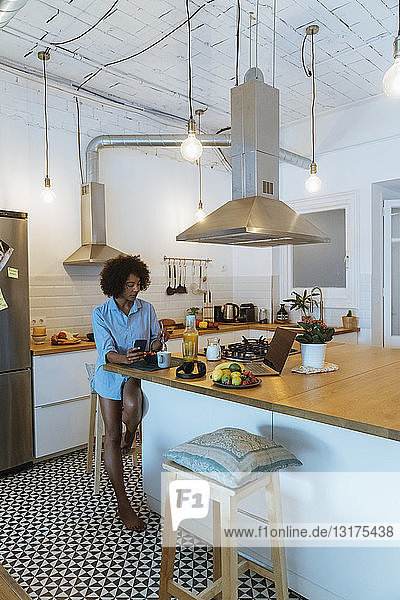 Frau frühstückt in ihrer Küche und benutzt ein Smartphone