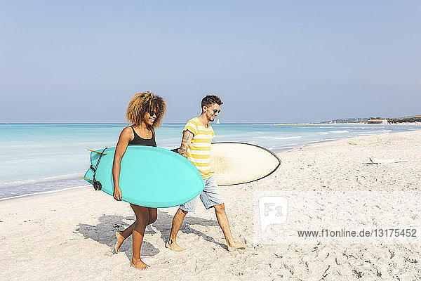 Paar  das am Strand spazieren geht und Surfbretter trägt