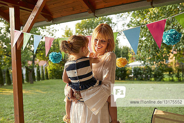 Glückliche Mutter trägt ihre Tochter auf einer Gartengeburtstagsfeier