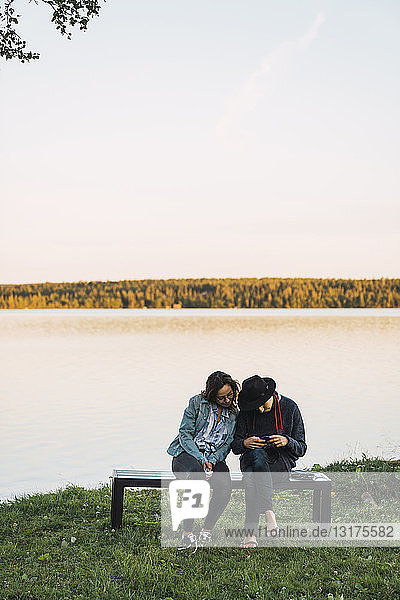 Junges Paar sitzt auf einer Bank am See und schaut sich die Bilder mit einer Kamera an