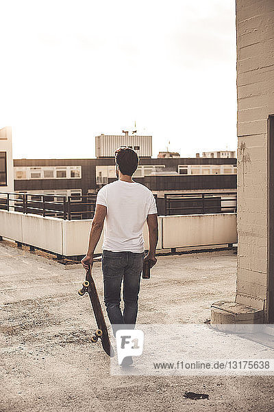 Rückansicht eines jungen Mannes mit Skateboard und Bierflasche auf der Dachterrasse in der Abenddämmerung