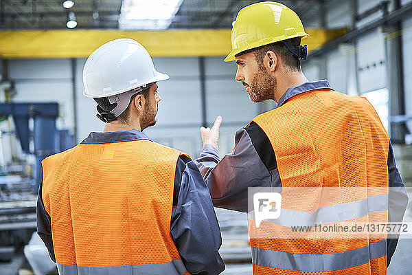 Rückansicht von zwei Männern in Arbeitsschutzkleidung  die sich in der Fabrik unterhalten