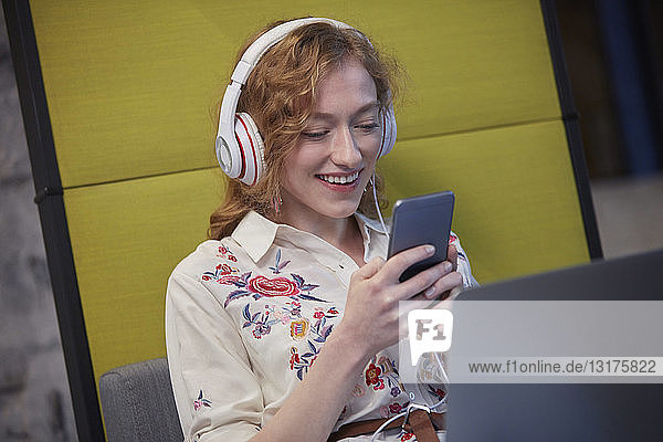 Junge Frau  die in einem modernen Kreativbüro arbeitet  Kopfhörer trägt und ein Smartphone benutzt