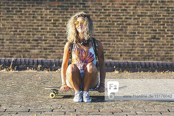 Junge Frau sitzt auf einem Skateboard mit Backsteinmauer im Hintergrund