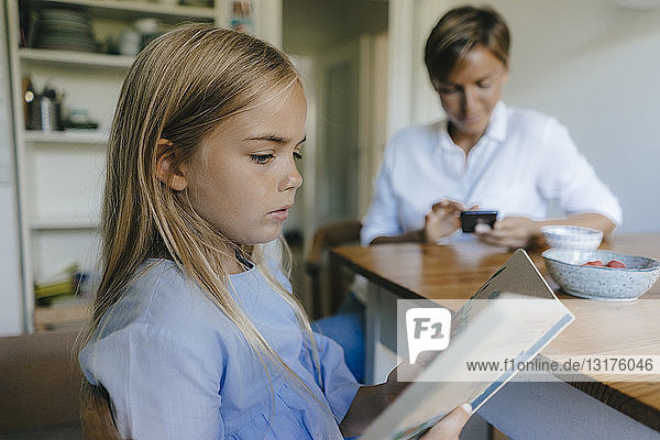 Girl with book sitting at table at home with mother using cell phone in background
