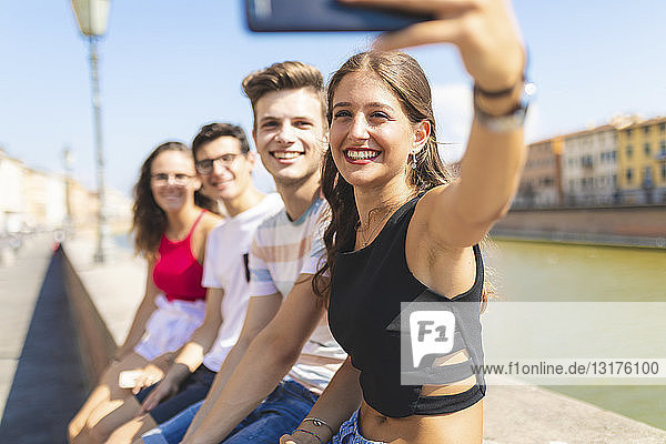 Italien,  Pisa,  eine Gruppe von vier glücklichen Freunden sitzt auf einer Mauer entlang des Arno und macht ein Selfie