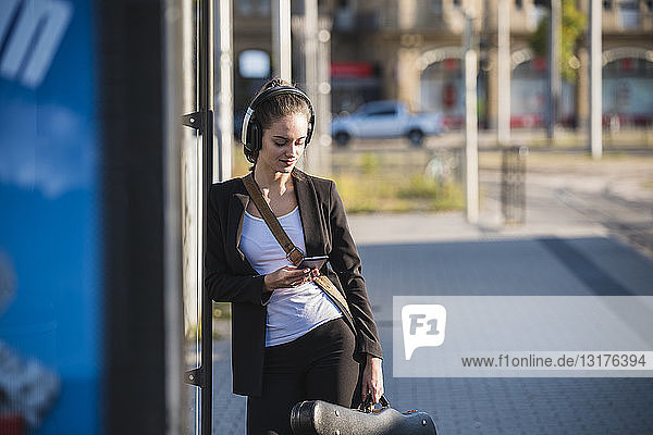 Junge Frau mit Kopfhörern und Handy an der Straßenbahnhaltestelle