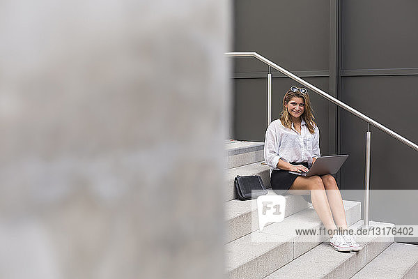 Lächelnde Geschäftsfrau sitzt mit Laptop auf der Treppe