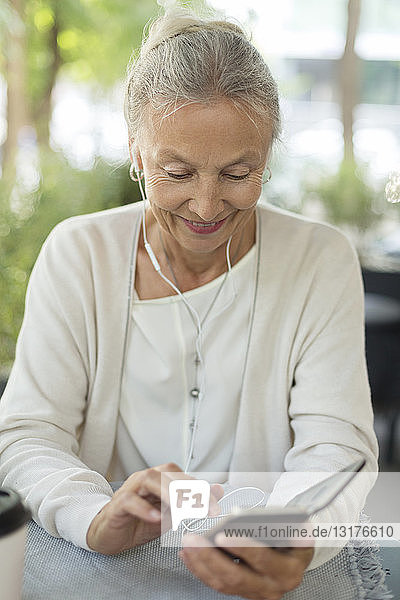 Lächelnde ältere Frau in einem Straßencafé mit Handy und Kopfhörern