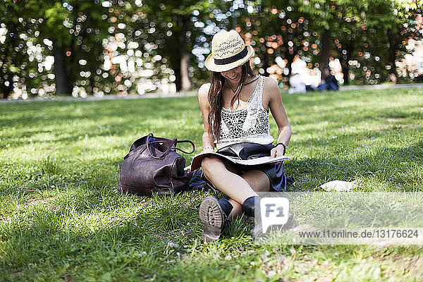 Lächelnder junger Student sitzt auf einer Wiese in einem Park und benutzt ein Heft