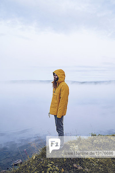 Schweden  Lappland  Mann mit Vollbart und gelber Windjacke steht am Wasser und schaut in die Ferne