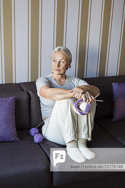 Porträt einer nachdenklichen älteren Frau  die mit Strickzeug auf der Couch sitzt und in die Ferne schaut