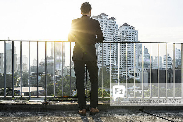 Business man in suit leaning on handrail in sunset on city rooftop