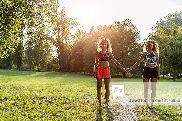 Zwillingsschwestern stehen Seite an Seite in einem Park im Gegenlicht und halten sich an den Händen