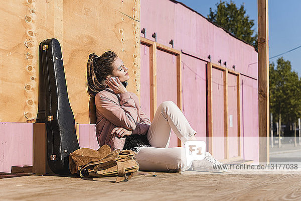 Lächelnde junge Frau sitzt auf dem Podest neben dem Gitarrenkoffer und hört Musik