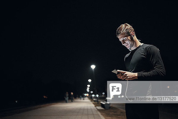 Sportlicher junger Mann mit Smartphone und Kopfhörern nachts im Freien