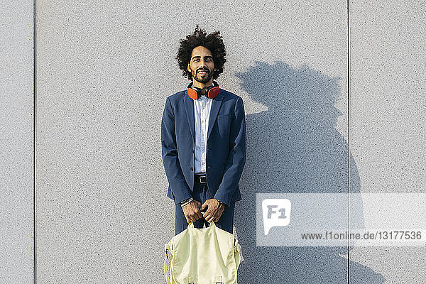 Porträt eines lächelnden jungen Geschäftsmannes mit Tasche und Kopfhörer an einer Wand