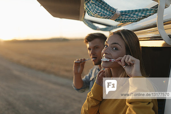 Ehepaar beim Zähneputzen am Wohnmobil in ländlicher Landschaft bei Sonnenuntergang