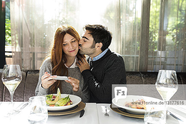 Man kissing woman taking a cell phone picture in a restaurant