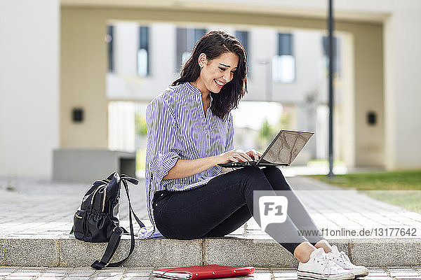 Smiling student sitting on stair outdoors working on laptop