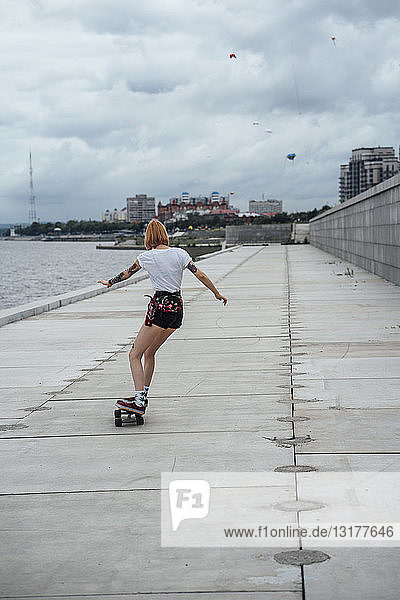 Rückansicht einer jungen Frau  die am Flussufer Carver-Skateboard fährt