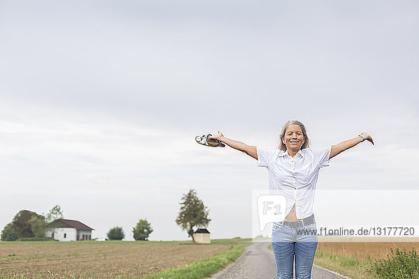Reife Frau steht im Sommer mit ausgestreckten Armen auf abgelegenem Feldweg