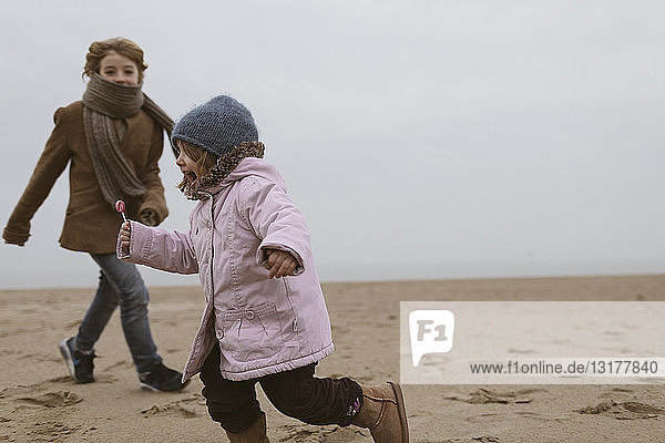 Schreiendes kleines Mädchen mit Lolly rennt im Winter am Strand herum  während ihr Bruder sie beobachtet