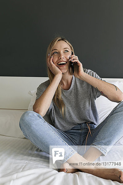 Happy young woman sitting on bed talking on cell phone