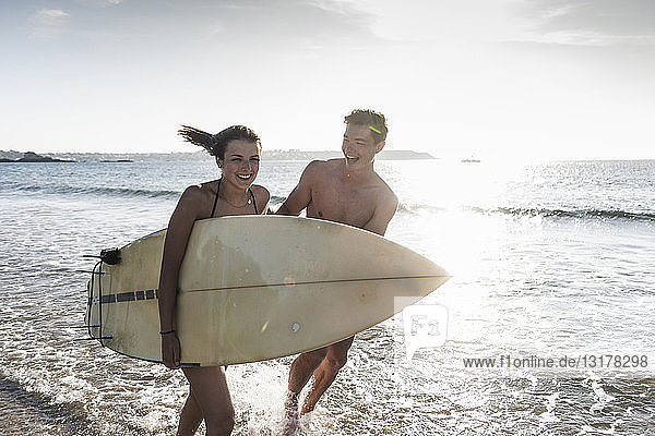 Frankreich  Bretagne  glückliches junges Paar mit im Meer laufendem Surfbrett