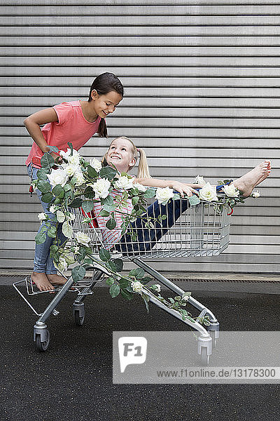 Zwei Mädchen spielen mit einem Einkaufswagen  der mit weißen Kunstblumen geschmückt ist