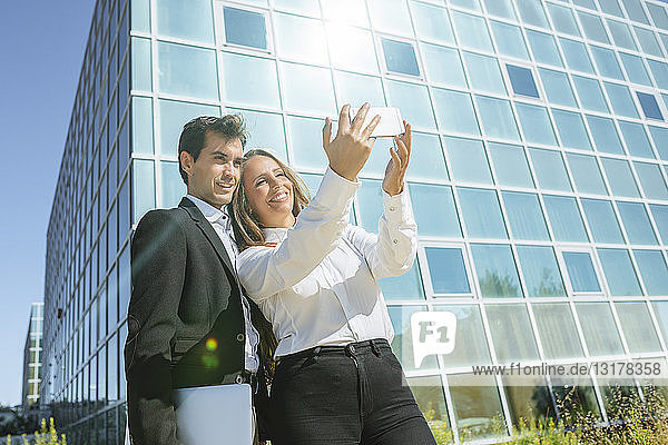 Lächelnde Geschäftsfrau und lächelnder Geschäftsmann beim Selfie vor dem Bürogebäude