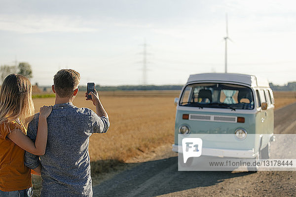 Young couple taking cell phone picture of camper van in rural landscape