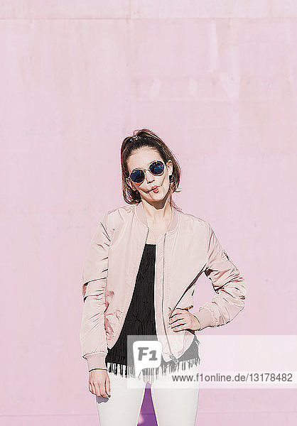Porträt einer jungen Frau mit Sonnenbrille  die vor einer rosa Wand grimassiert