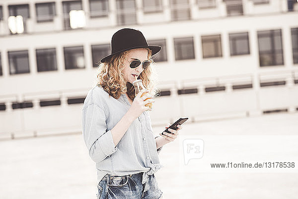 Junge Frau trinkt Getränk während sie ein Mobiltelefon benutzt