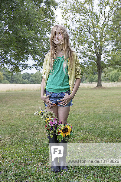 Portrait of blond girl standing on a meadow with bunches of flowers in her rubber boots