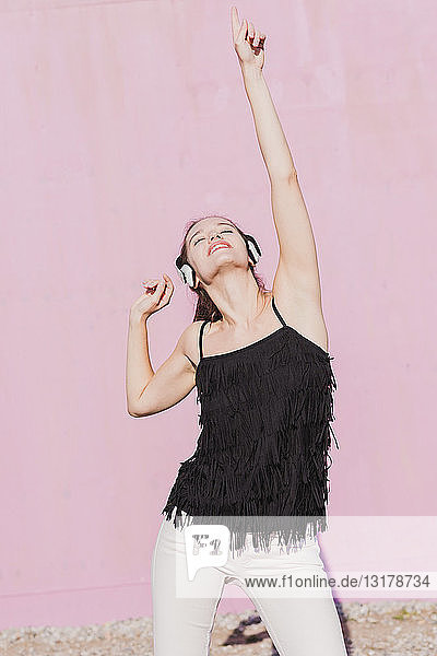 Glückliche junge Frau mit Kopfhörern tanzt vor rosa Wand