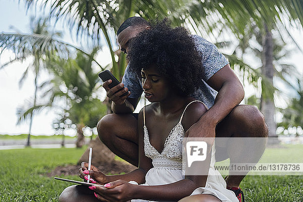 Junges Paar benutzt Tablet und Handy auf einem Rasen in einem Park