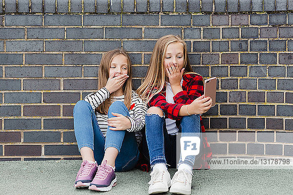 Porträt von zwei Mädchen  die vor einer Ziegelmauer sitzen und sich mit einem Smartphone selbstständig machen