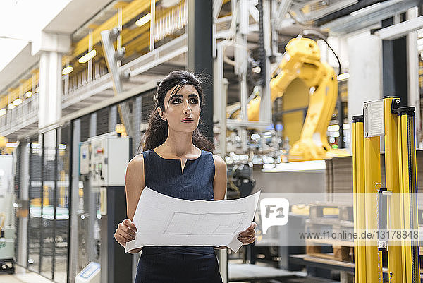 Frau hält Plan in Fabrikhalle mit Industrieroboter