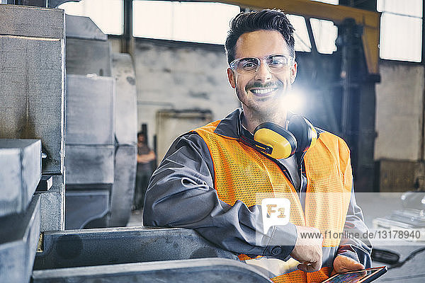 Porträt eines lächelnden Mannes  der Arbeitsschutzkleidung trägt und ein Tablett in der Fabrik hält