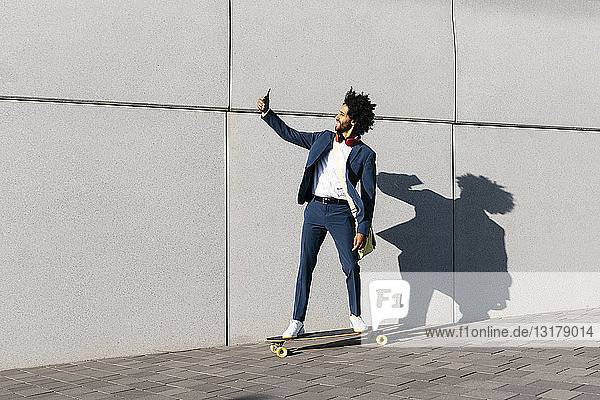 Junger Geschäftsmann fährt mit dem Skateboard an einer Wand entlang und nimmt einen Selfie