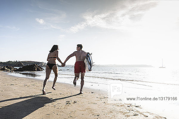 Frankreich, Bretagne, junges Paar mit einem Surfbrett, das Hand in Hand im Meer läuft