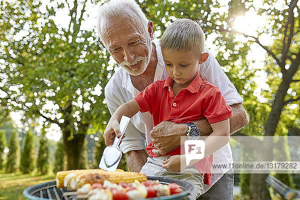 Großvater hilft Enkel beim Wenden eines Maiskolbens während eines Grillfestes im Garten