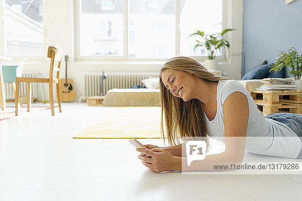 Lächelnde junge Frau liegt auf dem Boden in ihrem Loft und schaut auf ihr Handy